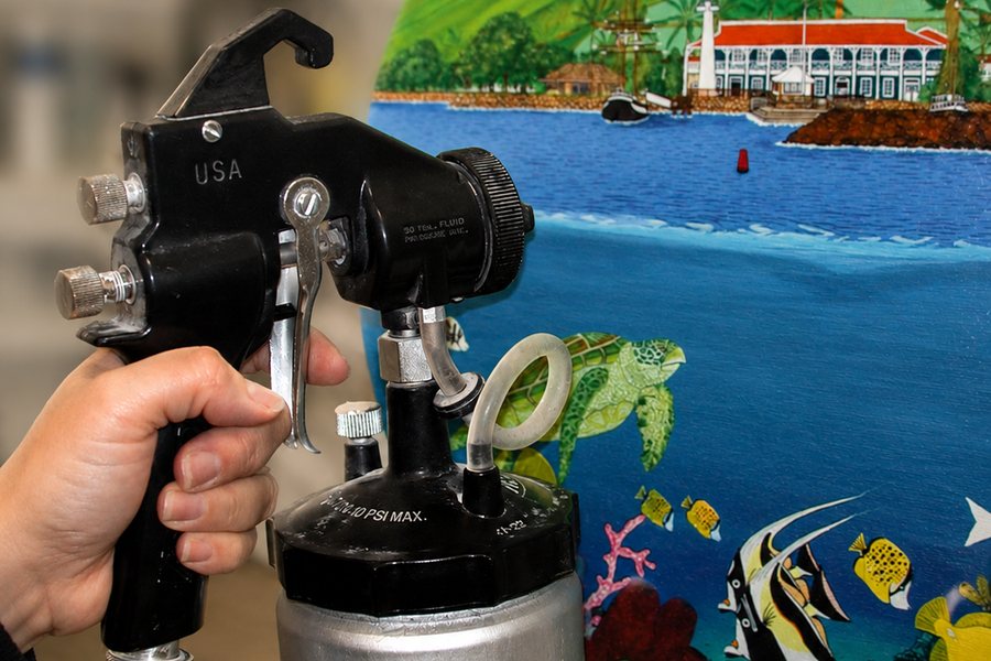 A spray gun applying a protective print varnish to a colorful painted harbour scene.